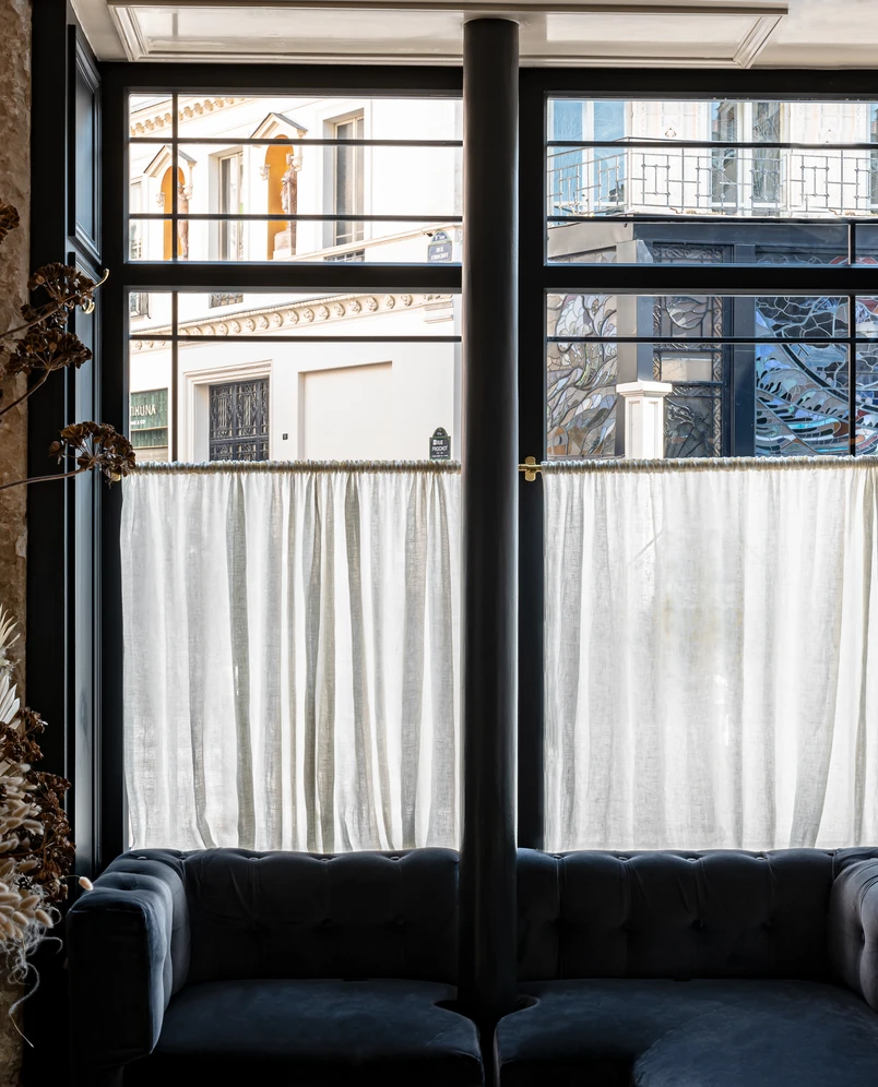 Interior view of a dark blue sofa by a large window with sheer white curtains partially covering the outside street scene.