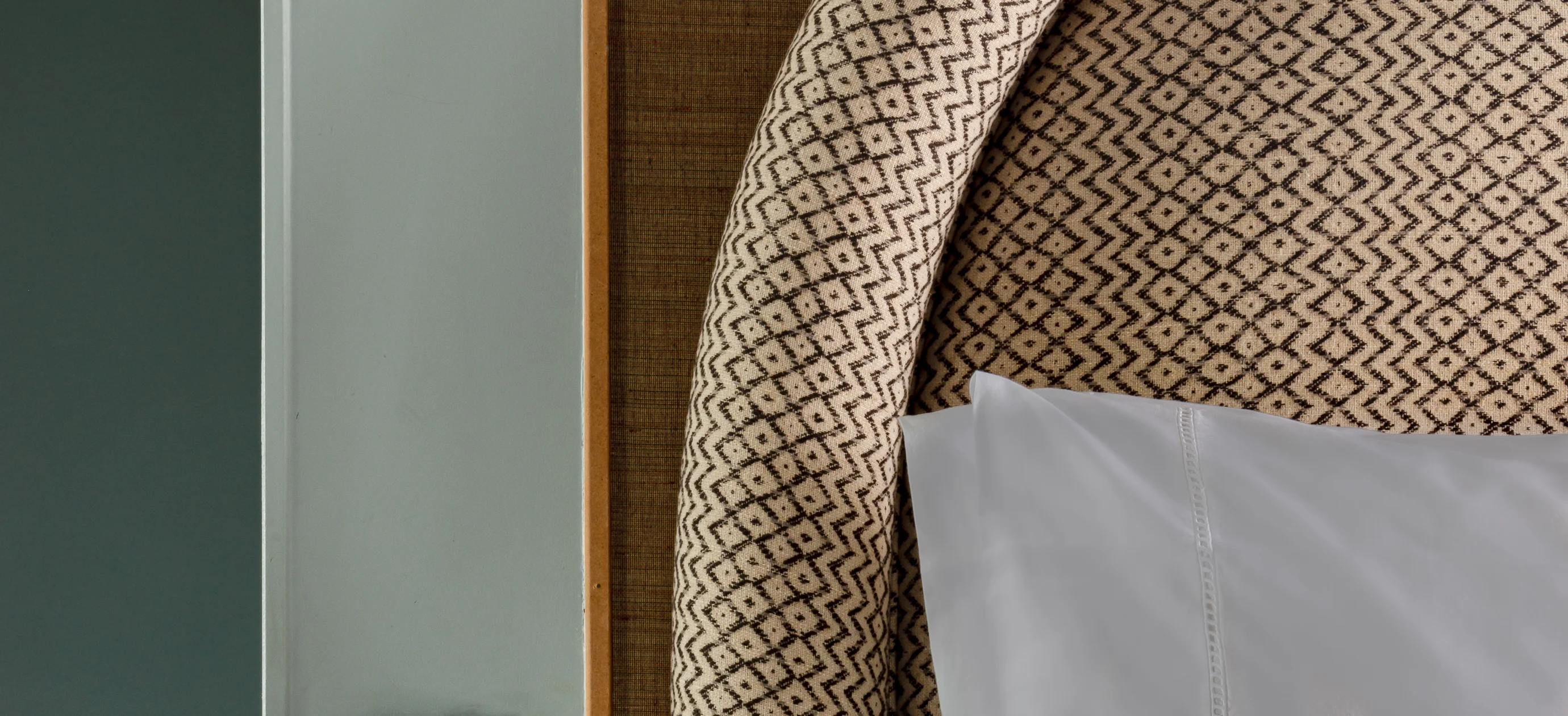 Close-up of a patterned upholstered headboard and white pillow on a bed beside a wall.