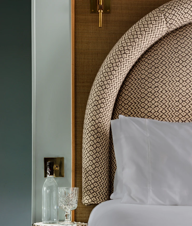 Close-up of a patterned upholstered headboard with a white pillow and bedsheet, beside a glass bottle and crystal glass on a table.