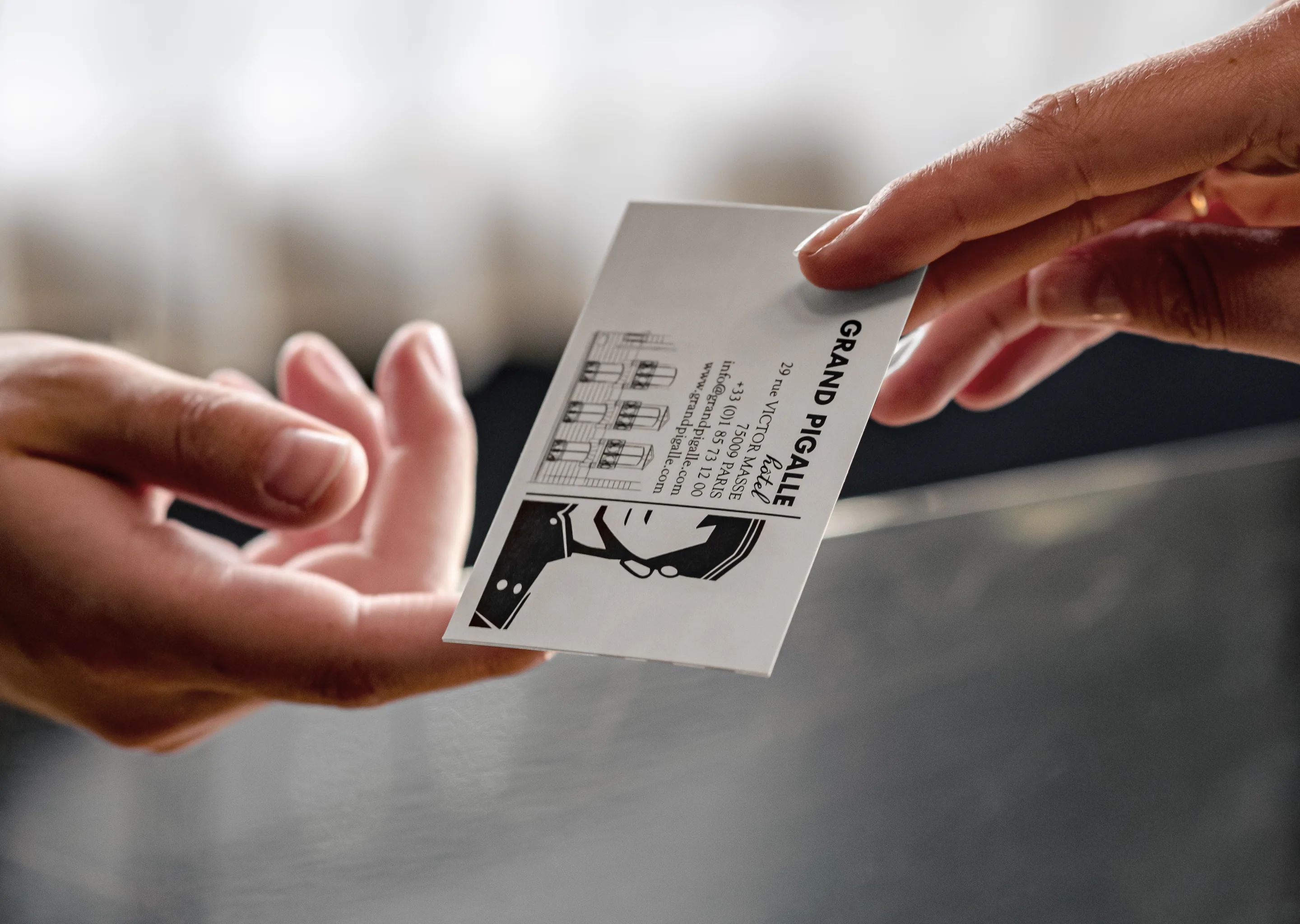 Two hands exchanging a Grand Pigalle hotel business card at a reception desk.