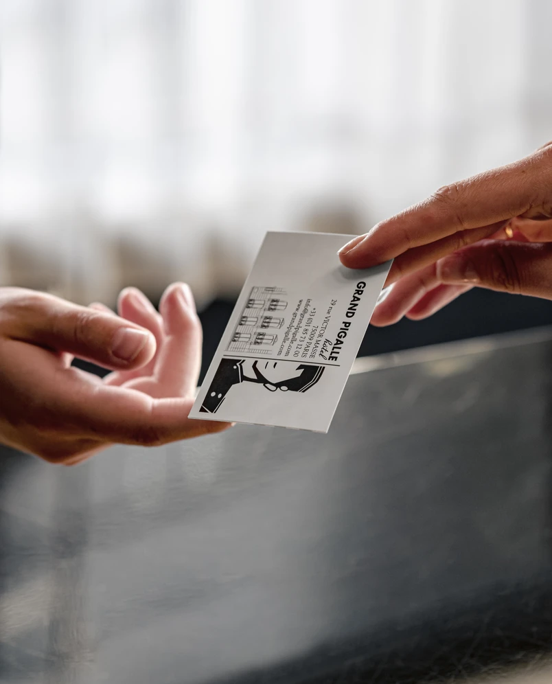 Close-up of one hand handing a Grand Pigalle hotel business card to another hand over a dark surface.