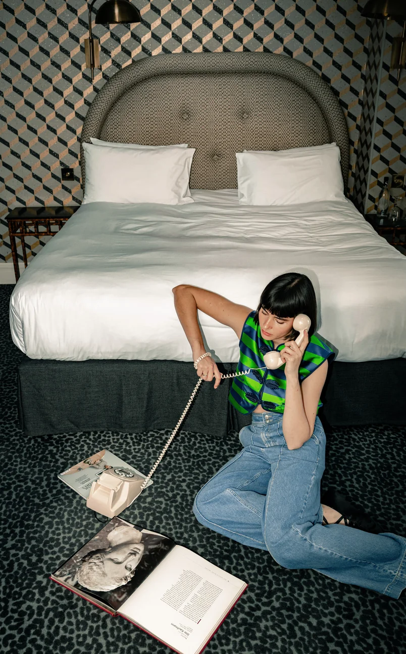 Woman with black bob haircut and sleeveless green and blue striped top sitting on leopard print carpet, talking on a vintage beige rotary phone in a hotel room.
