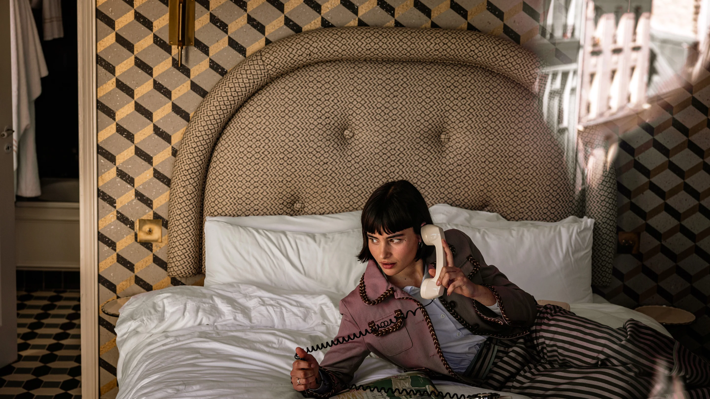 Mujer de pelo corto y oscuro con una chaqueta estampada y pantalones a rayas acostada en una cama mientras sostiene un receptor de teléfono con cable blanco.