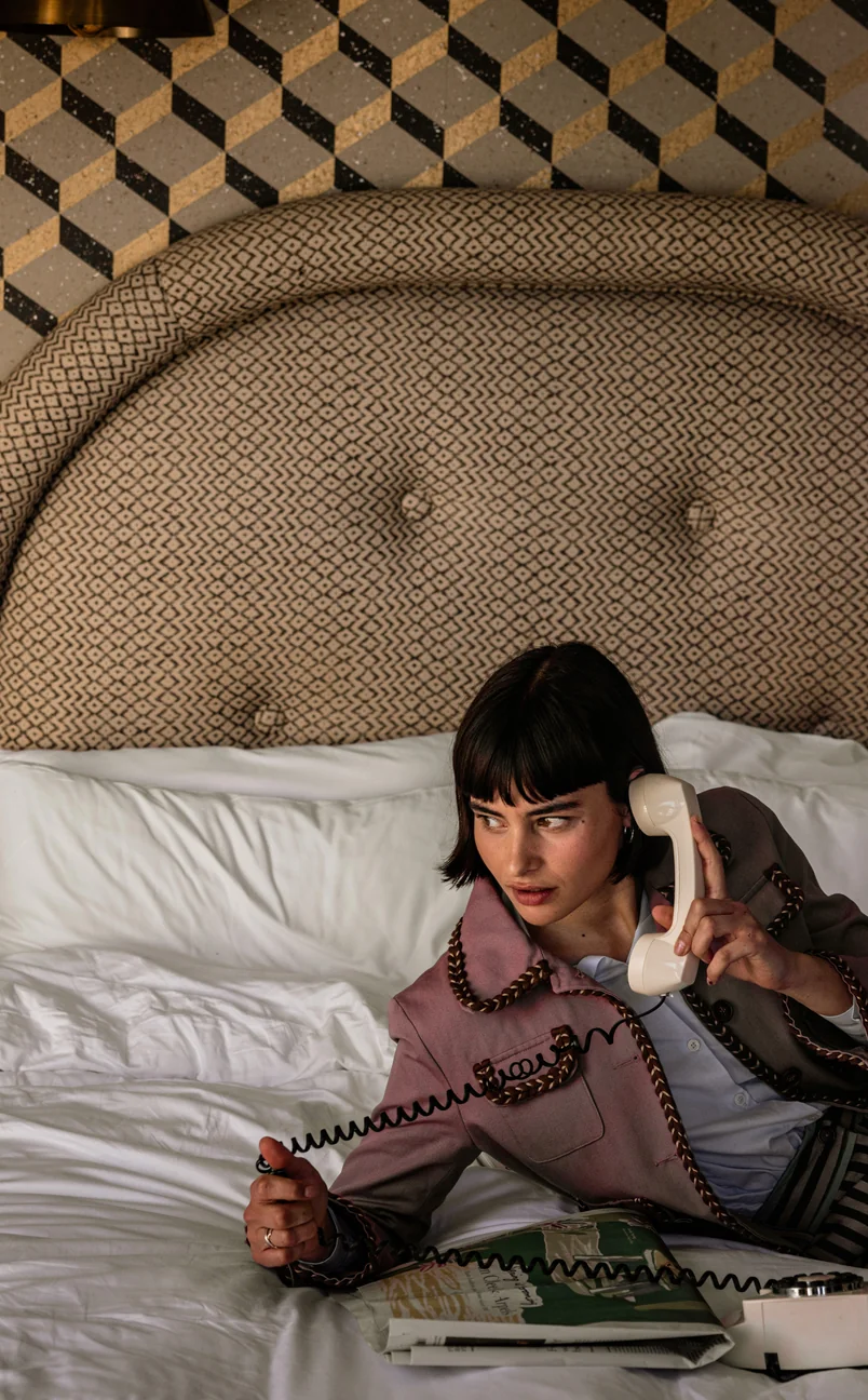 Mujer de pelo corto y oscuro acostada en una cama, sosteniendo un receptor de teléfono con cable de color crema, con una cabecera estampada y un fondo de pared de azulejos.