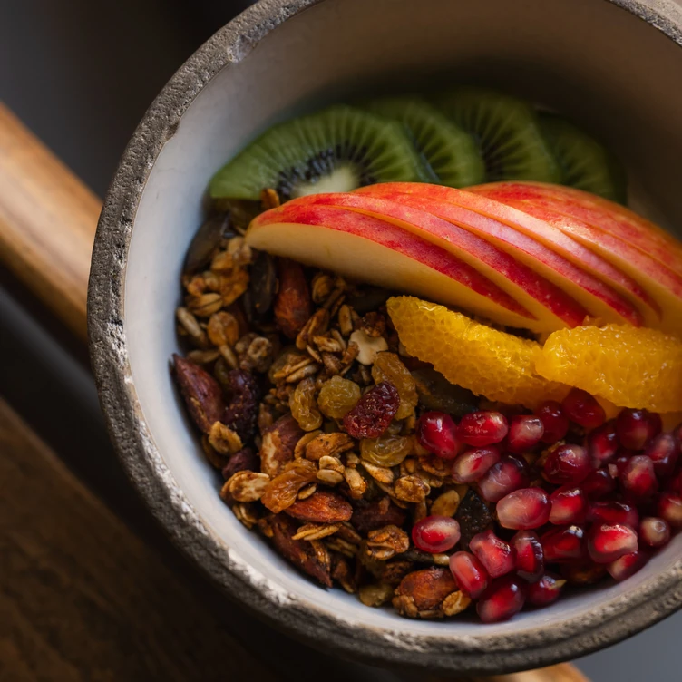 Bowl of granola topped with slices of apple, kiwi, orange segments, and pomegranate seeds.
