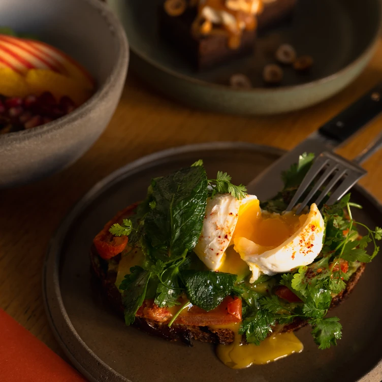 Slice of toast topped with melted cheese, fresh greens, and a poached egg with runny yolk on a dark plate.