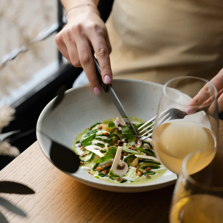 Person cutting a gourmet dish with green sauce, mushrooms, and garnishes in a bowl, accompanied by a glass of white wine on a wooden table.