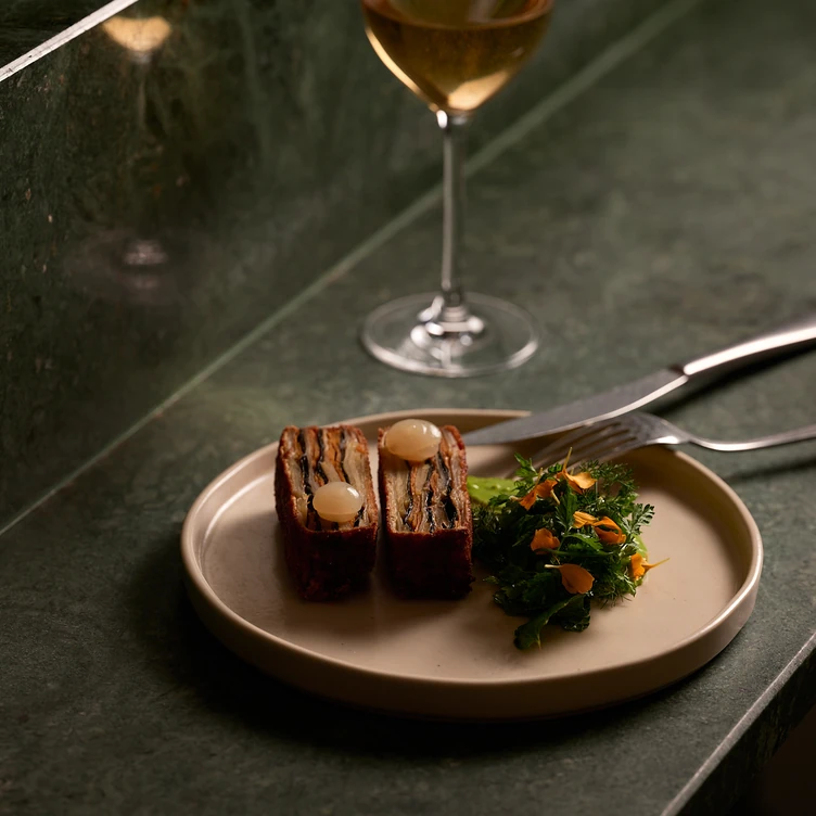 Two gourmet layered slices topped with pearl onions served with a small salad on a beige plate next to a glass of white wine.