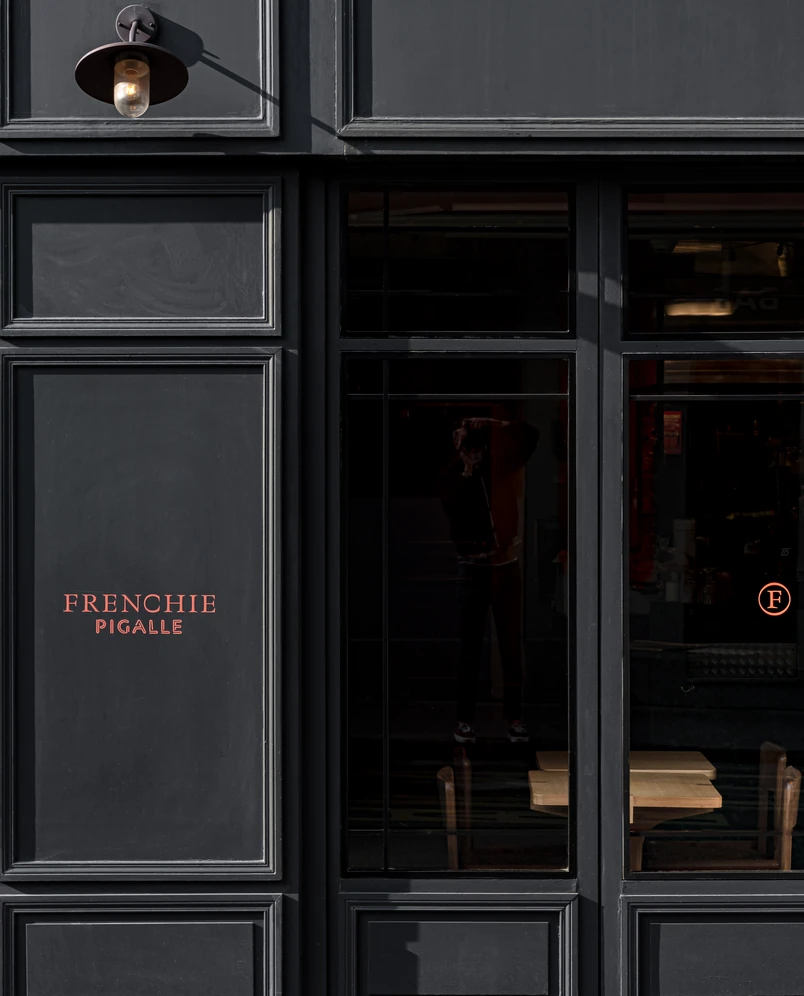 Black facade of Frenchie Pigalle restaurant with orange text and reflection in glass door showing wooden tables inside.