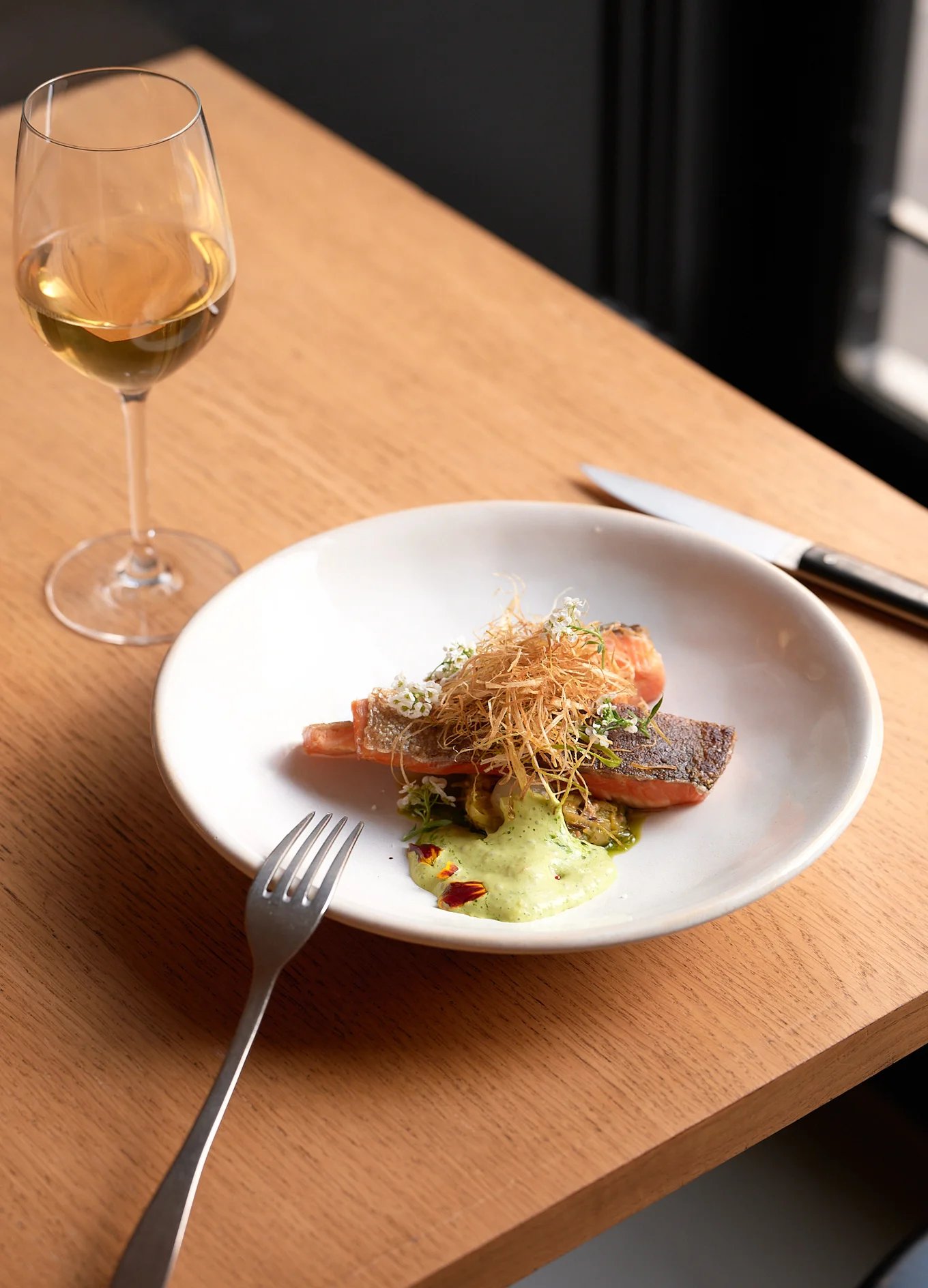 Plated dish of cooked salmon topped with crispy garnish and herbs, accompanied by a green sauce, with a glass of white wine on the side.