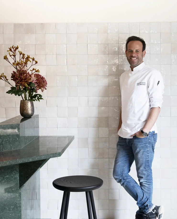 Smiling man in a white chef jacket and jeans standing with hands in pockets against a white tiled wall next to a marble countertop with a vase of flowers and a black stool.