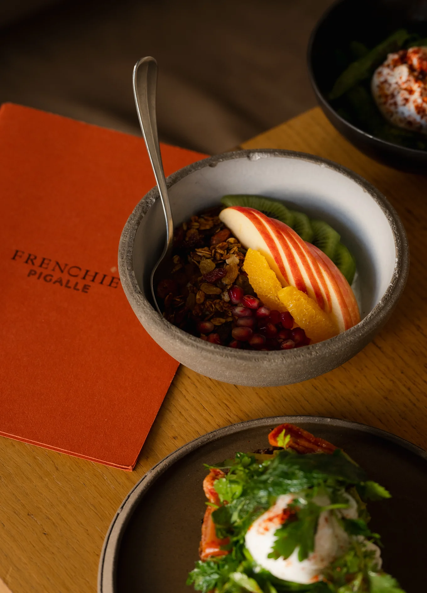 Bowl of granola topped with sliced apple, kiwi, orange, and pomegranate seeds on a wooden table next to a Frenchie Pigalle menu and a plate with a salad and poached egg.