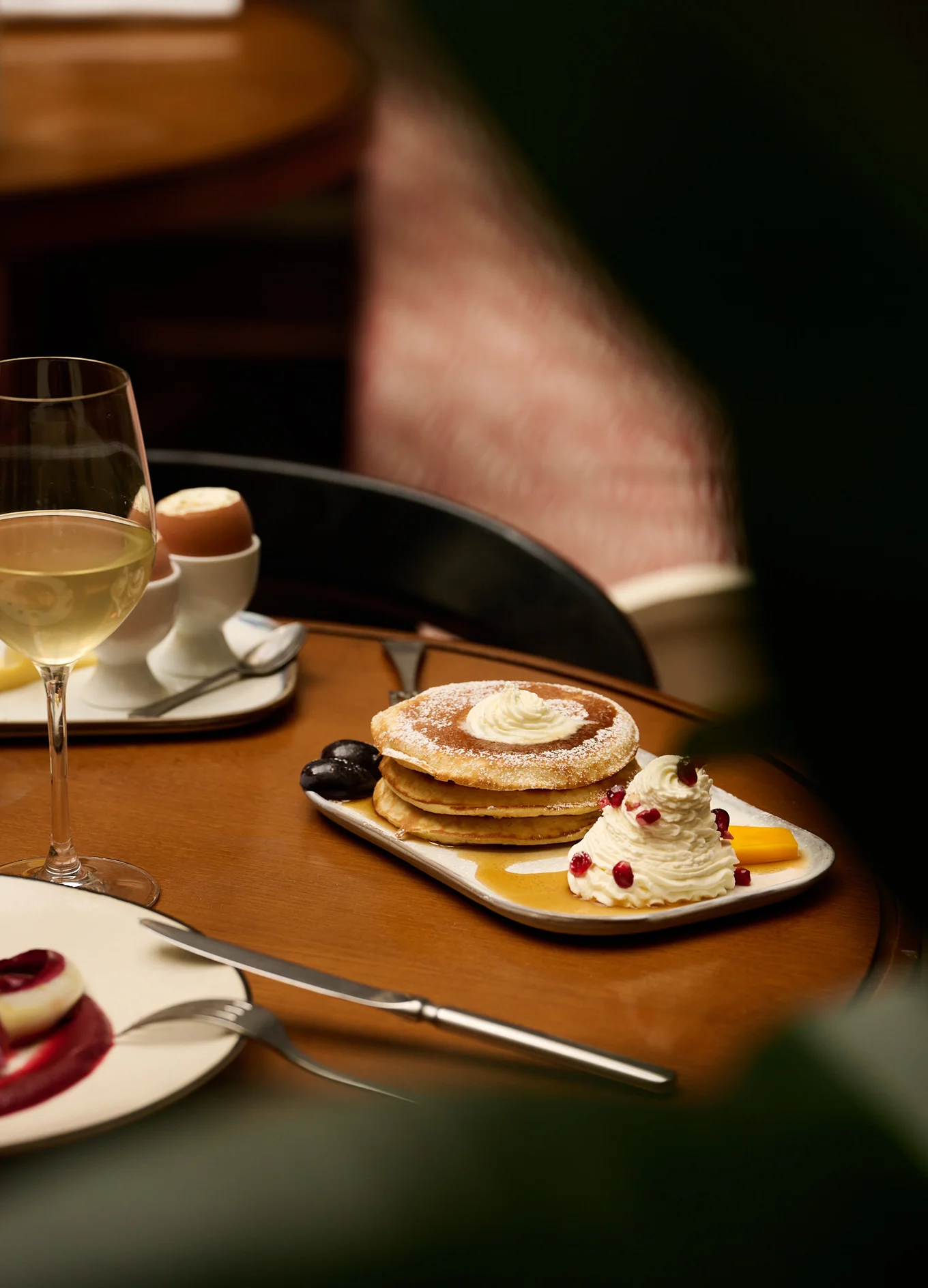 Stack of pancakes with whipped cream, syrup, pomegranate seeds, and a glass of white wine on a wooden table.