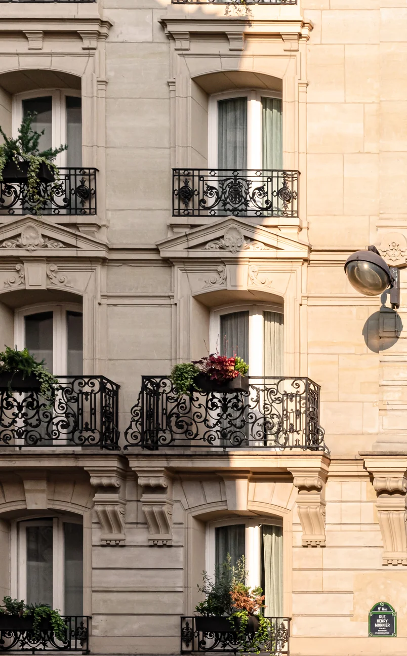 Primer plano de la fachada de un edificio de piedra de color beige con balcones ornamentados de hierro forjado negro adornados con macetas y una farola vintage en el lado derecho.