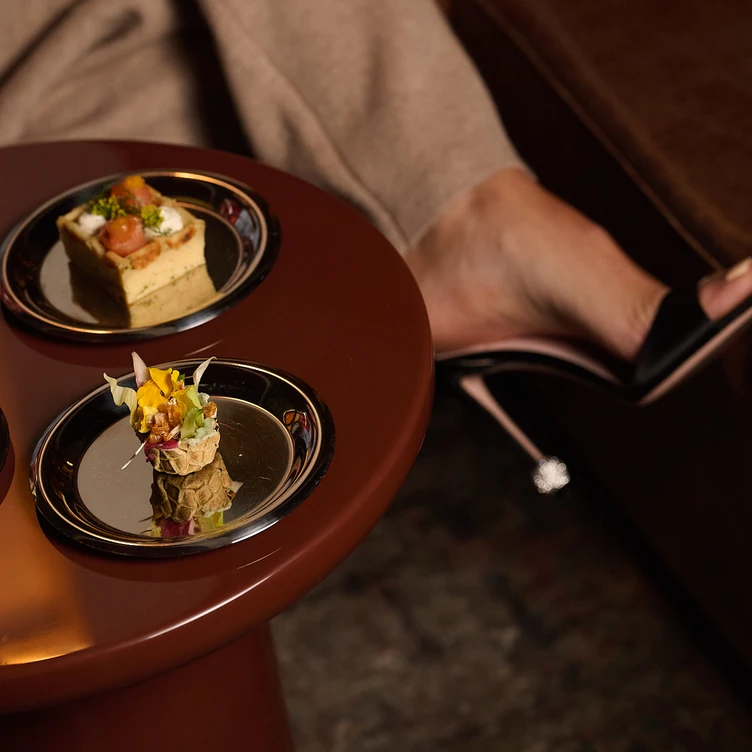 Dos pequeños aperitivos servidos sobre una mesa con una persona con pantalones beige y tacones negros de fondo.