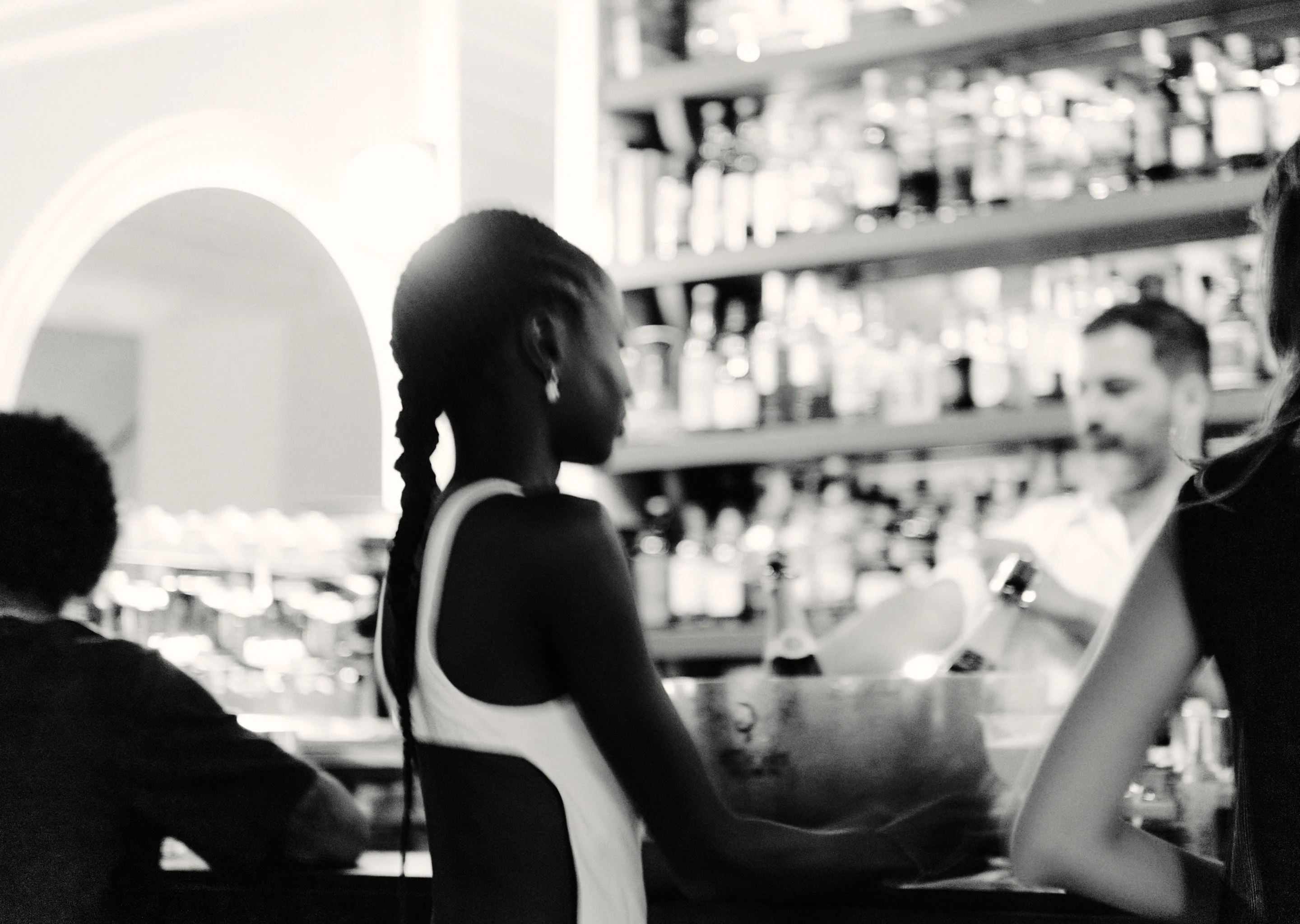 Foto en blanco y negro de una mujer con el pelo trenzado de pie en la barra de un bar con botellas de fondo.