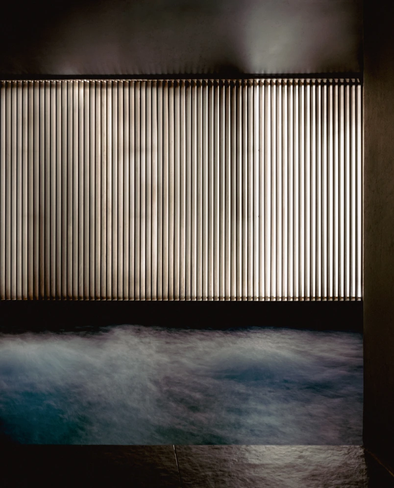 Indoor pool with bubbling water beneath a large window covered by vertical blinds with light shining through.
