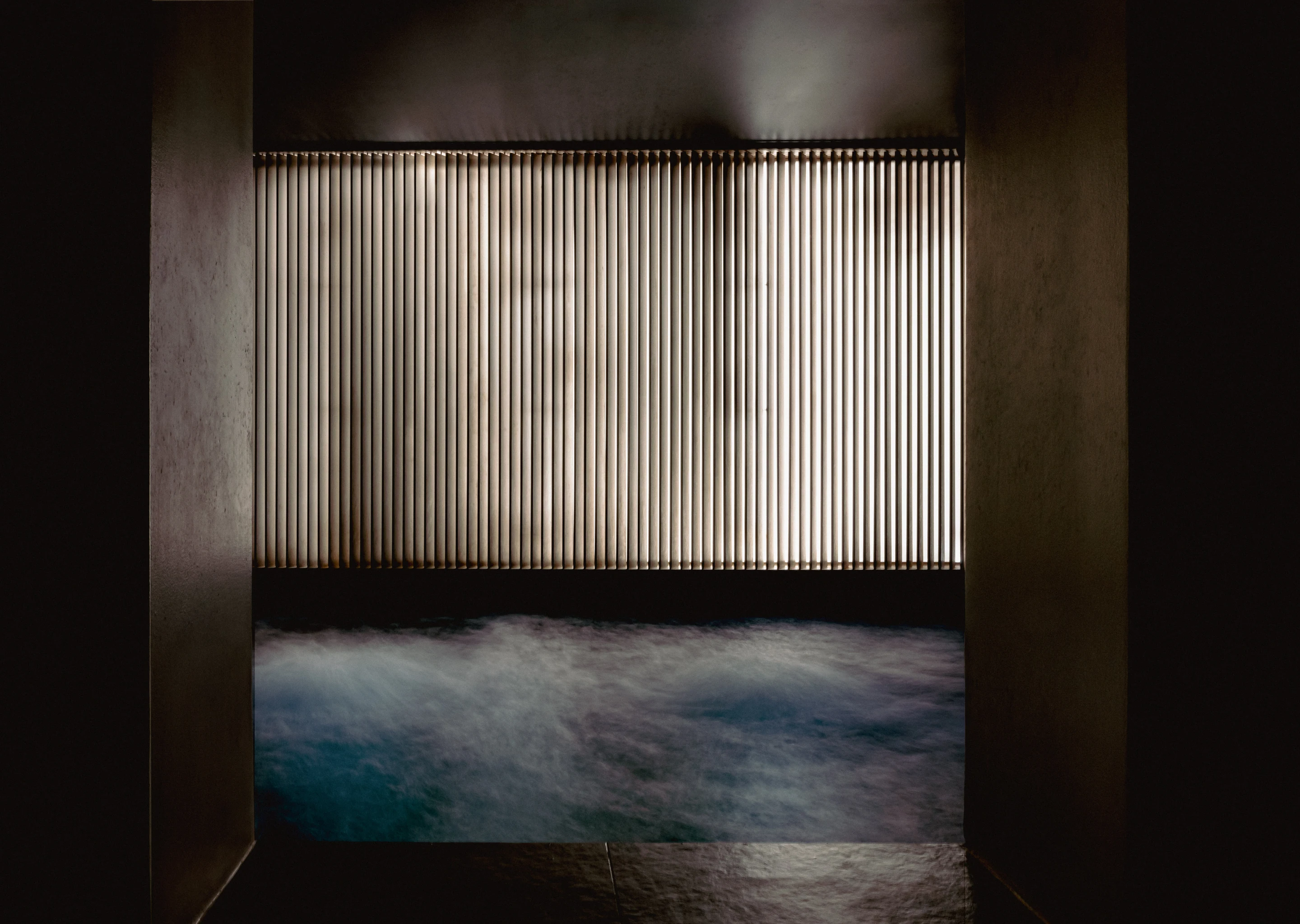 Indoor spa pool with bubbling water and a vertical slat illuminated wall in a dark room.