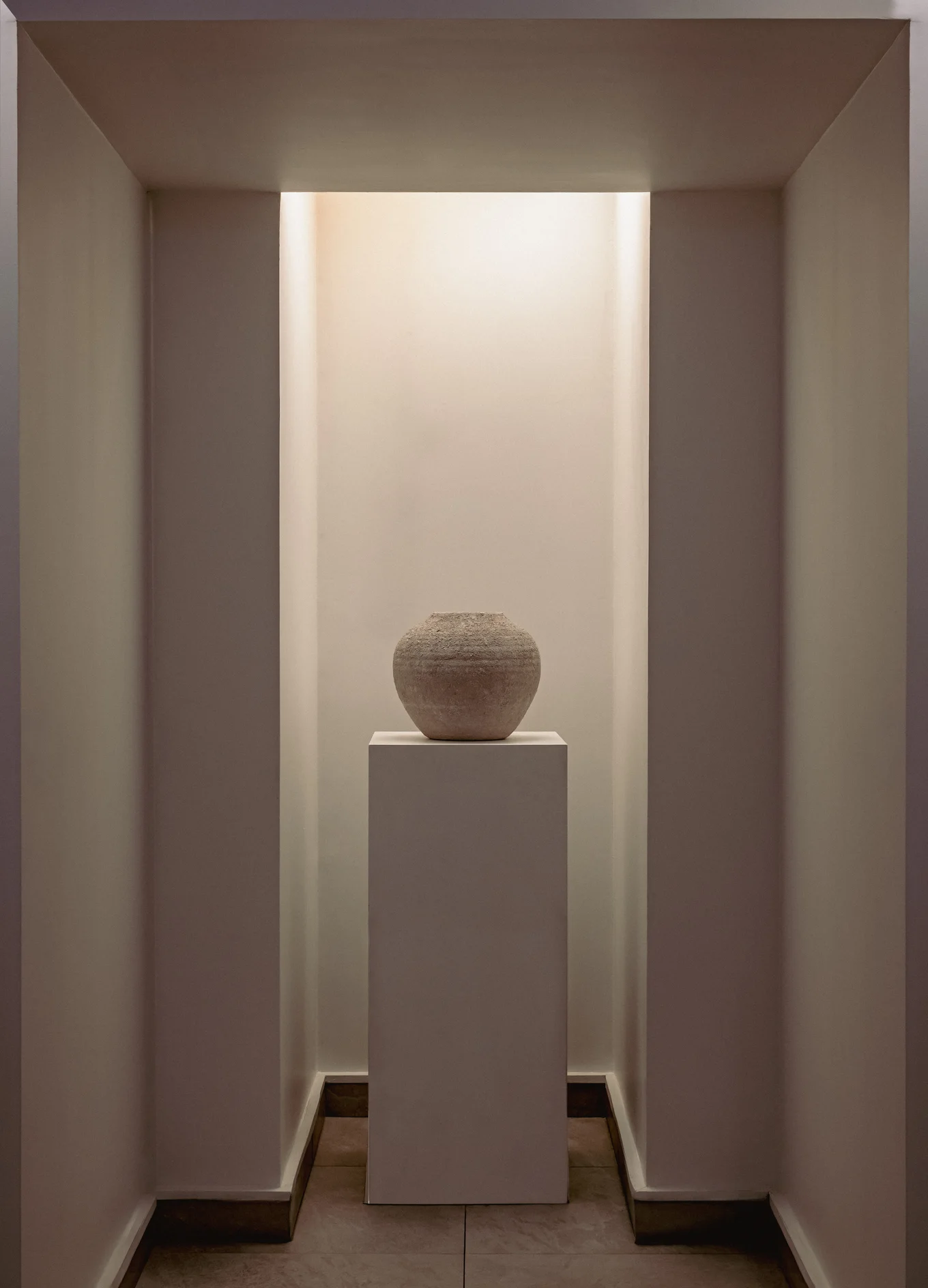 Rounded stone vase displayed on a white pedestal in a softly lit, minimalist alcove.