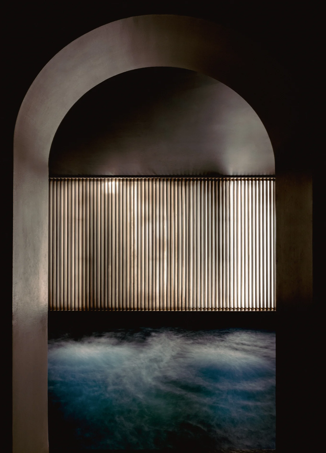 Dark spa area with a softly lit vertical slatted wall and water gently rippling in the foreground.
