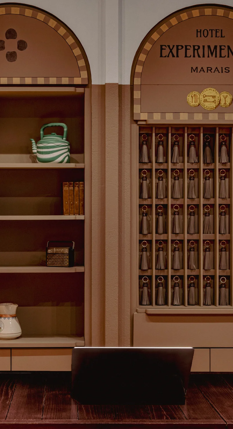 Reception area of Hotel Experimental Marais with shelves holding a green and white teapot, books, a radio, and a rack of keychains with tassels.