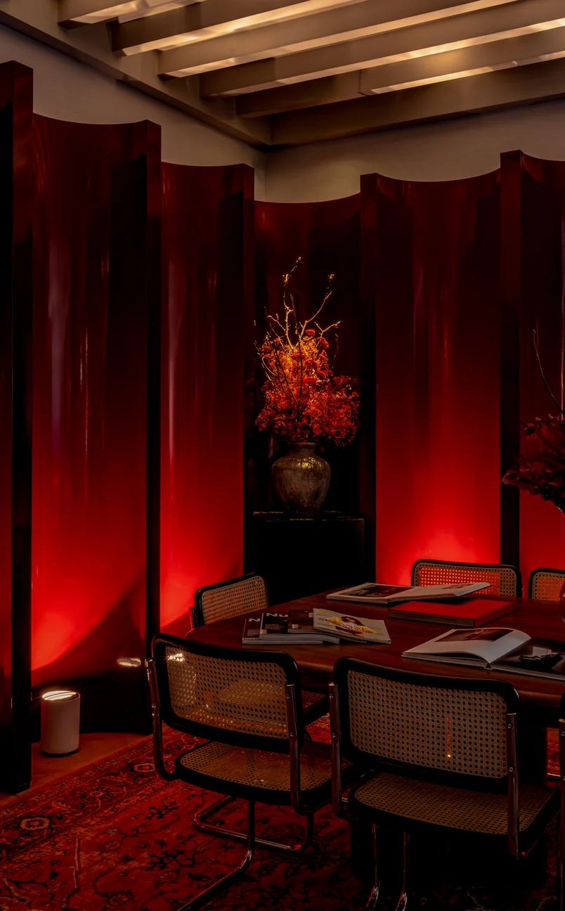 Dimly lit room with dark wood paneling, a vase of orange flowers, a wooden table with magazines, and woven cane chairs on a red patterned rug.