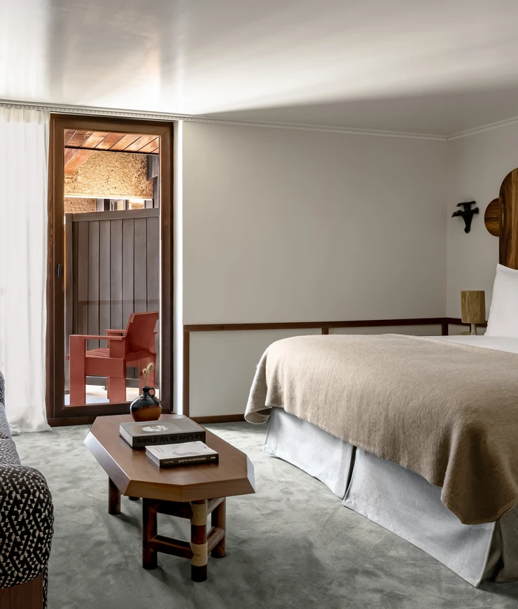 Minimalist bedroom with a beige bedspread, wooden coffee table with books and vase, and a glass door opening to a balcony with a red chair.