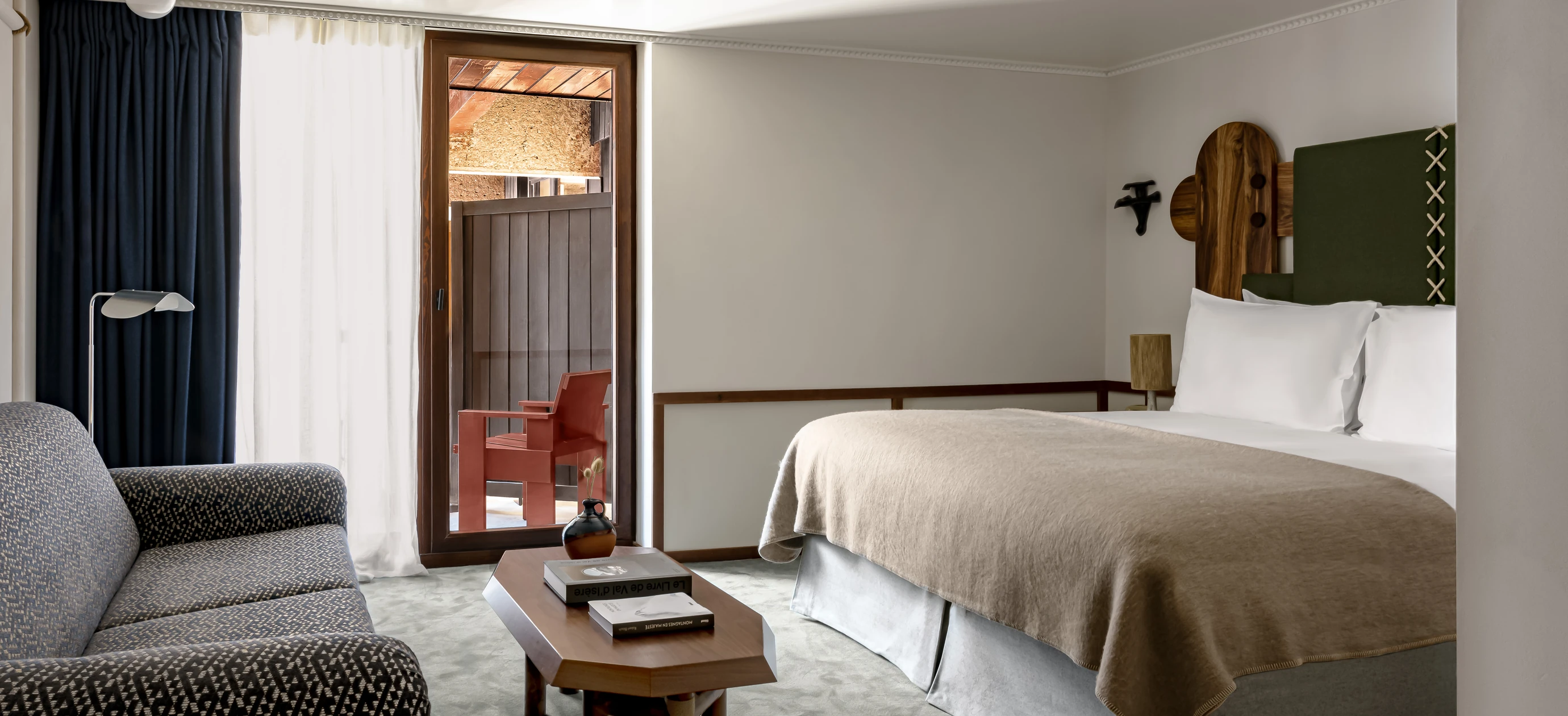 Hotel room with a large bed, patterned sofa, wooden table with books and vase, and a balcony with a red chair.