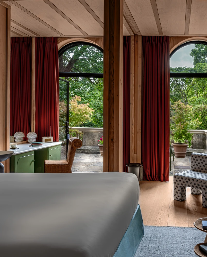 Hotel room with a bed, green cabinet, wicker chair, and open arched doors leading to a terrace with potted plants and greenery outside.