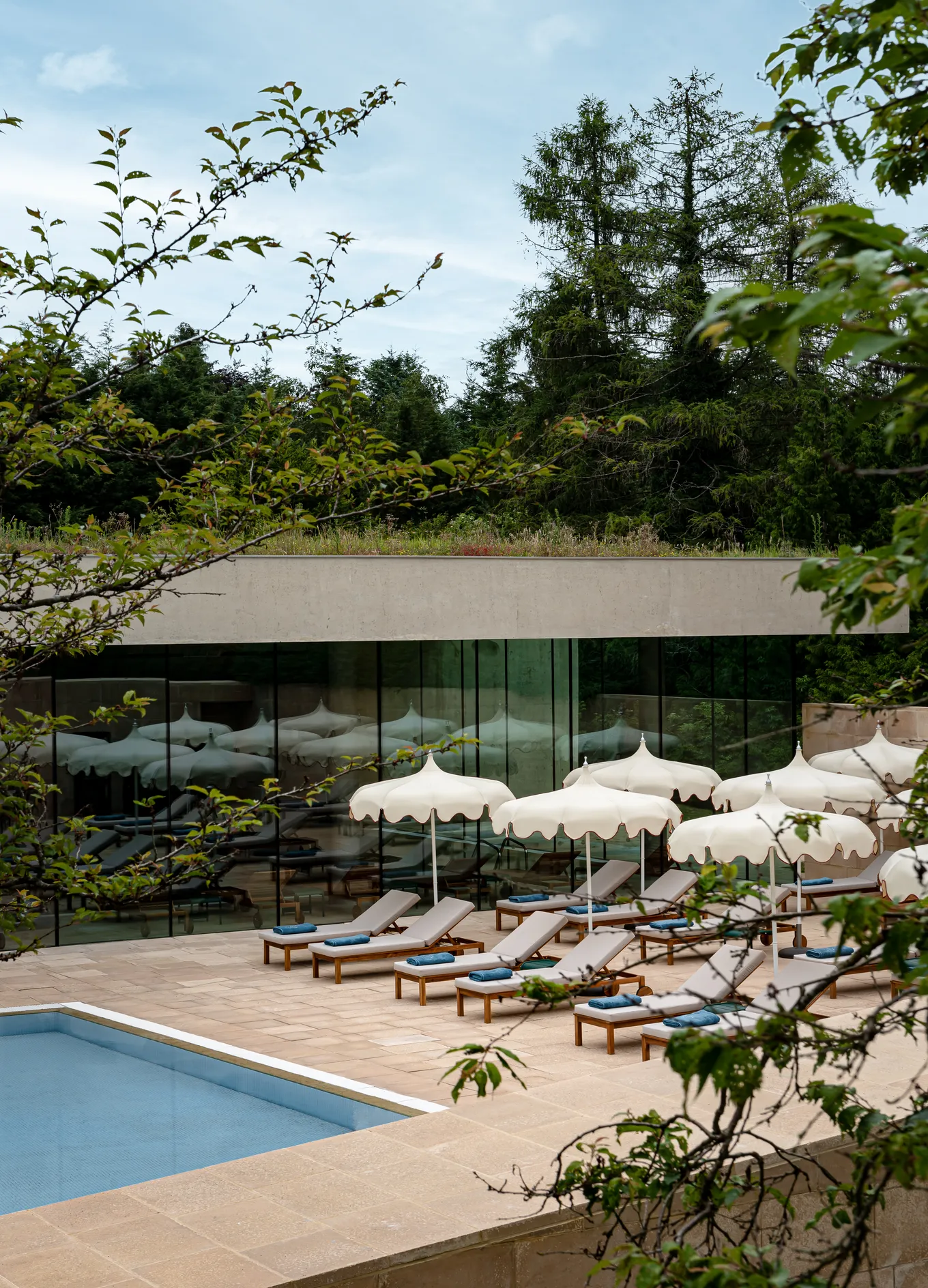 Outdoor pool area with beige lounge chairs and white parasols in front of a modern glass building surrounded by trees.