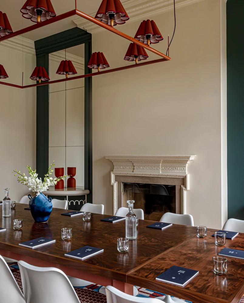 Sala de conferências elegante com uma longa mesa de madeira, cadeiras brancas, vasos azuis com flores, copos, cadernos e luzes vermelhas refletidas em um espelho perto de uma lareira decorativa.