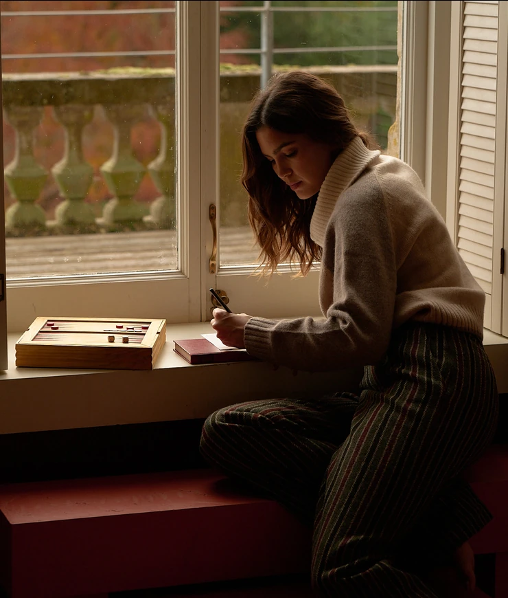 Woman in a cozy sweater writing in a notebook while sitting on a window bench with a backgammon game nearby.