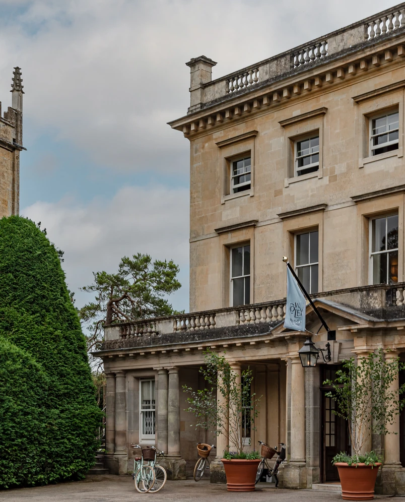 Historic stone building with columns and a Flag reading 'Cowley Manor', bicycles parked near entrance, and large green bushes.