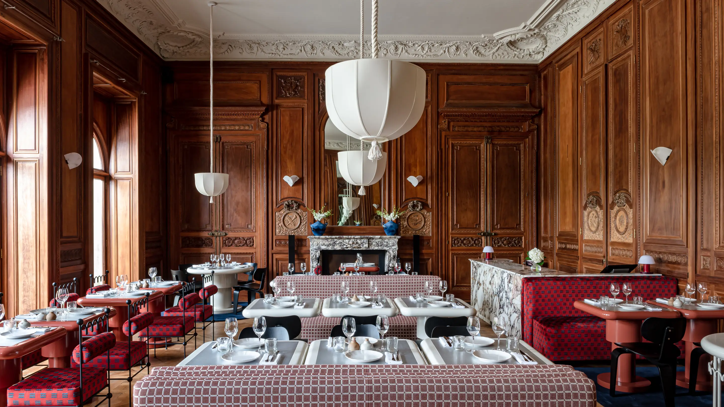 Elegant restaurant interior with ornate wooden wall paneling, marble fireplace, white pendant lights, and red patterned seating.