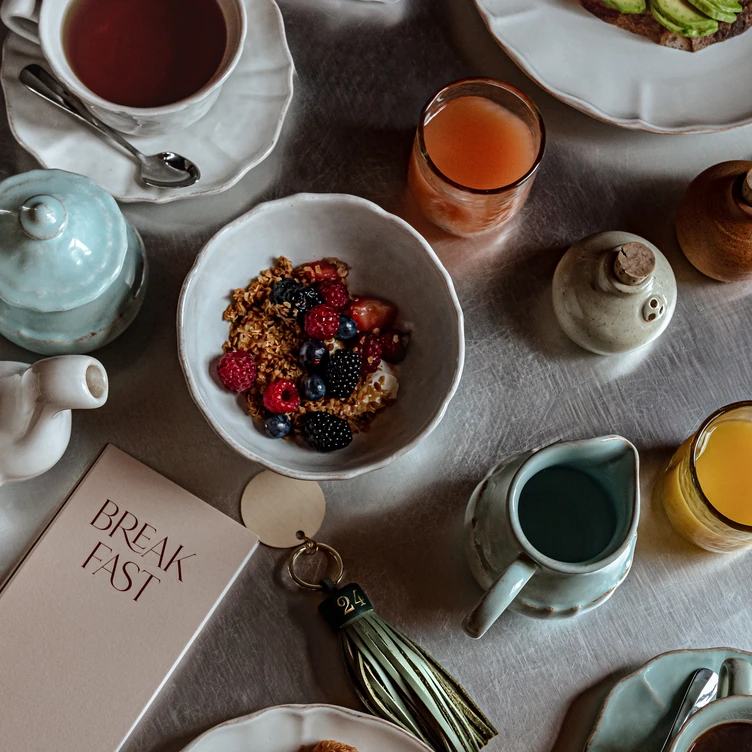 Table setting with a bowl of granola and berries, tea in a cup, two glasses of juice, ceramic pitchers, salt and pepper shakers, and a menu card labeled 'BREAKFAST'.