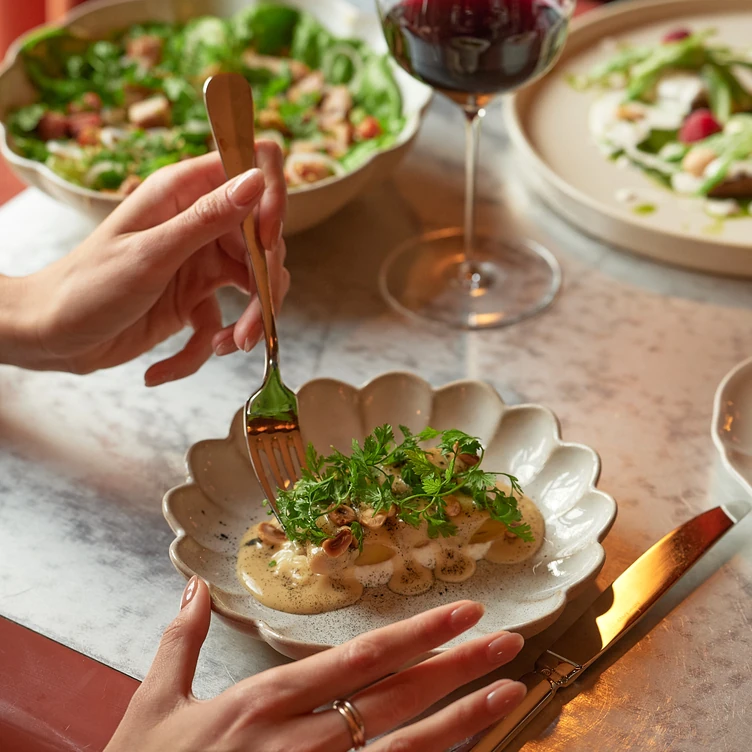 Person using a fork to eat a gourmet dish with creamy sauce, herbs, and nuts on a scalloped plate with a glass of red wine and salad bowls in the background.