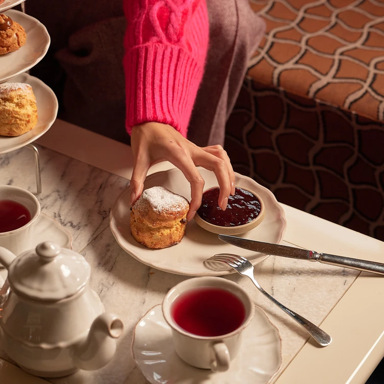 Hand in a pink sweater reaching for a powdered scone on a plate with jam, next to cups of tea and a white teapot on a marble table.