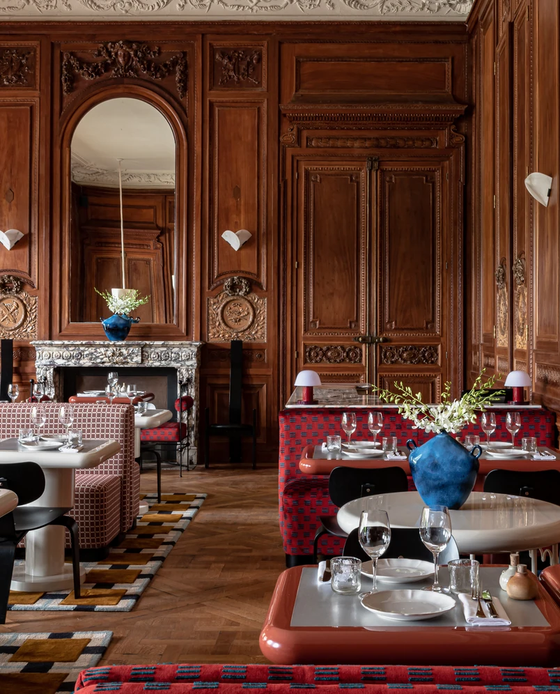 Elegant dining room with carved wooden paneling, ornate ceiling, patterned red and beige chairs, and tables set with glassware and white plates, featuring blue vases with white flowers.