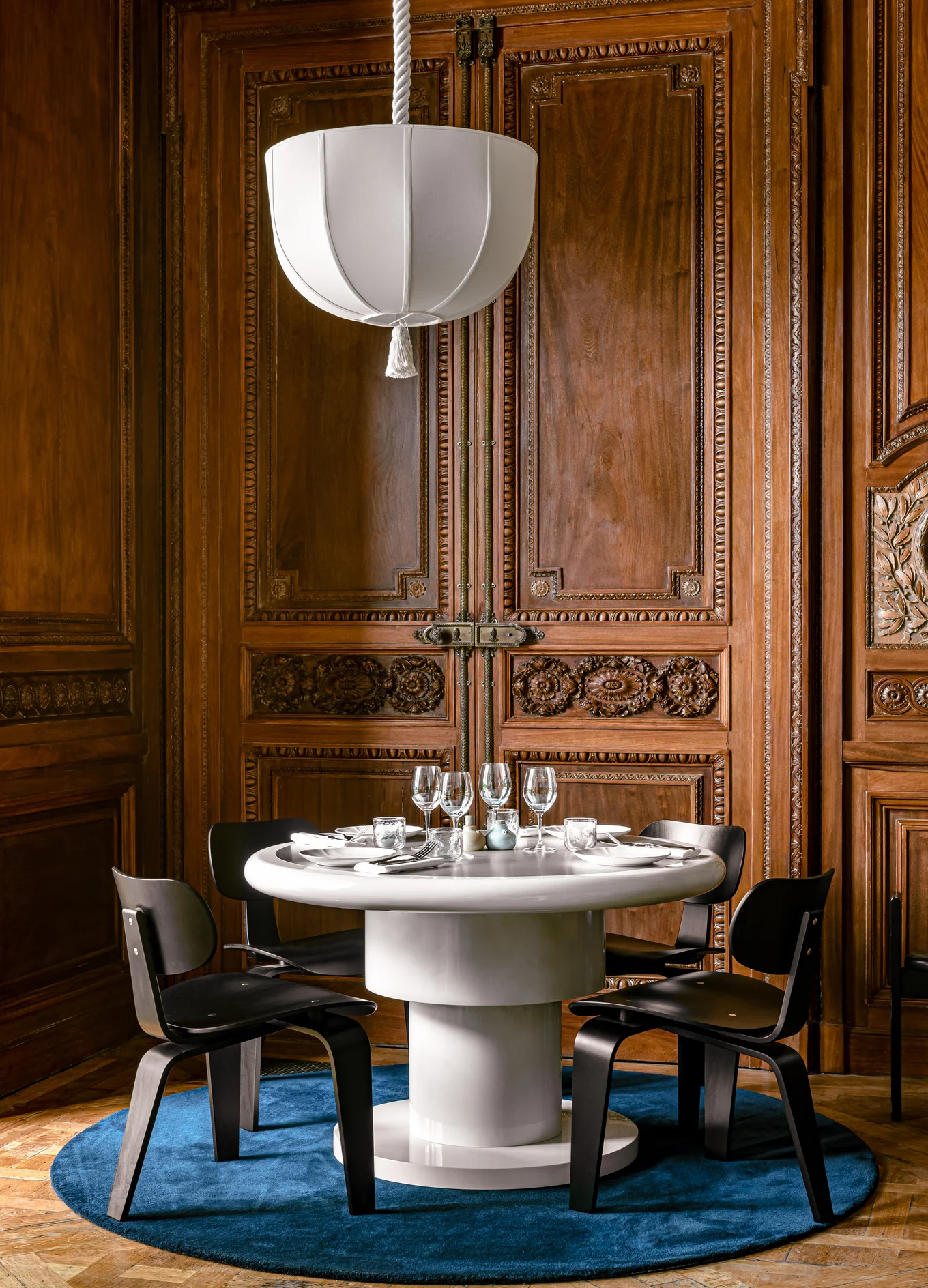 Round white dining table set with glassware and plates, surrounded by black chairs on a blue rug in front of ornately carved wooden doors, under a white pendant lamp.