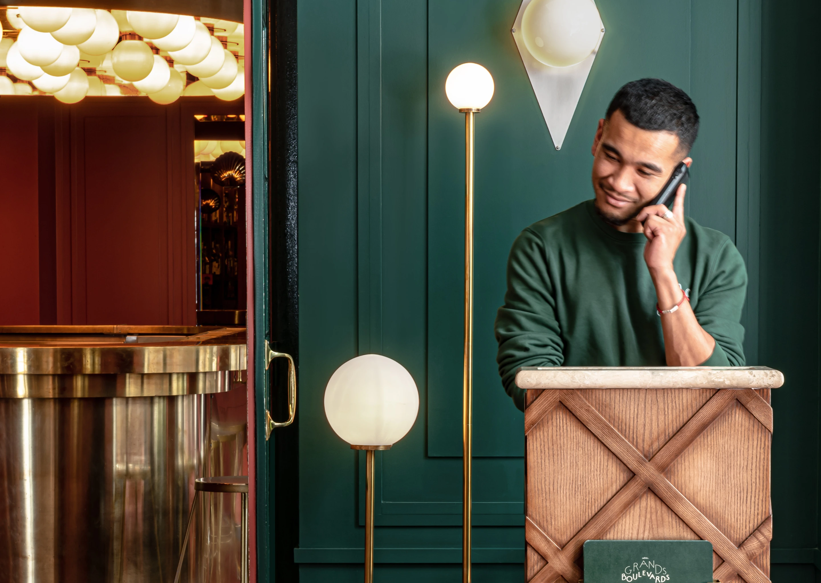 Man in a green sweater talking on a phone behind a wooden reception desk in a stylish restaurant interior with green walls and brass lighting.