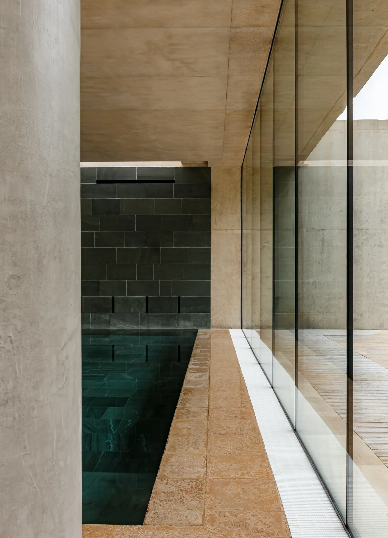 Modern indoor pool with dark tiled wall, beige stone floor, and large glass windows reflecting the ceiling.