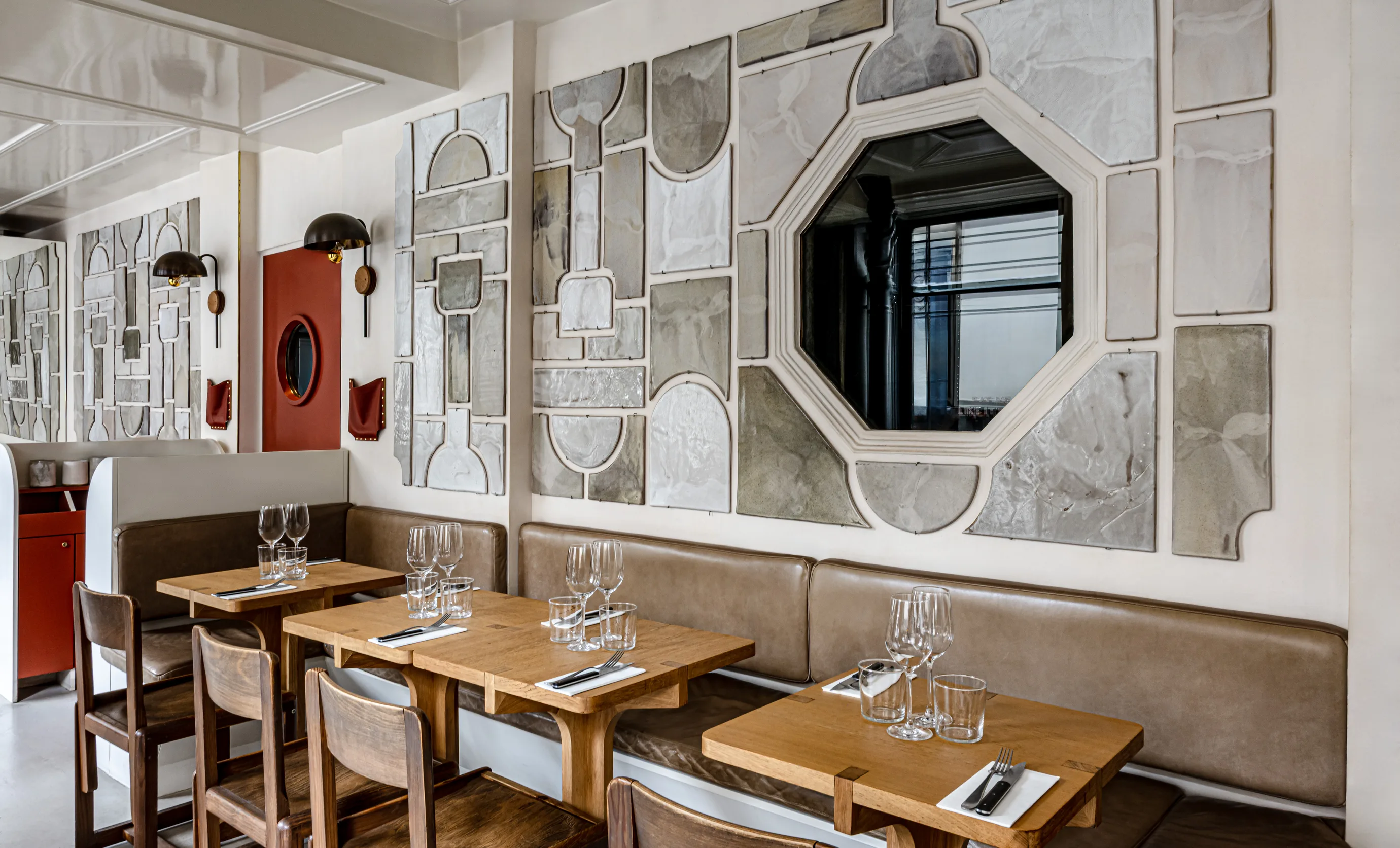 Modern restaurant interior with wooden tables, brown leather bench seating, wooden chairs, and abstract gray wall art including an octagonal mirror.