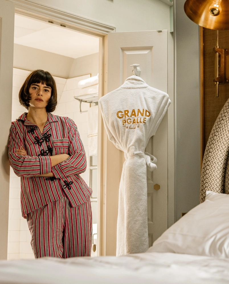 Mujer en pijama de rayas rojas y blancas apoyada en el marco de una puerta en una habitación de hotel, con un albornoz blanco colgado en la puerta bordado con la inscripción «GRAND PIGALLE hotel».