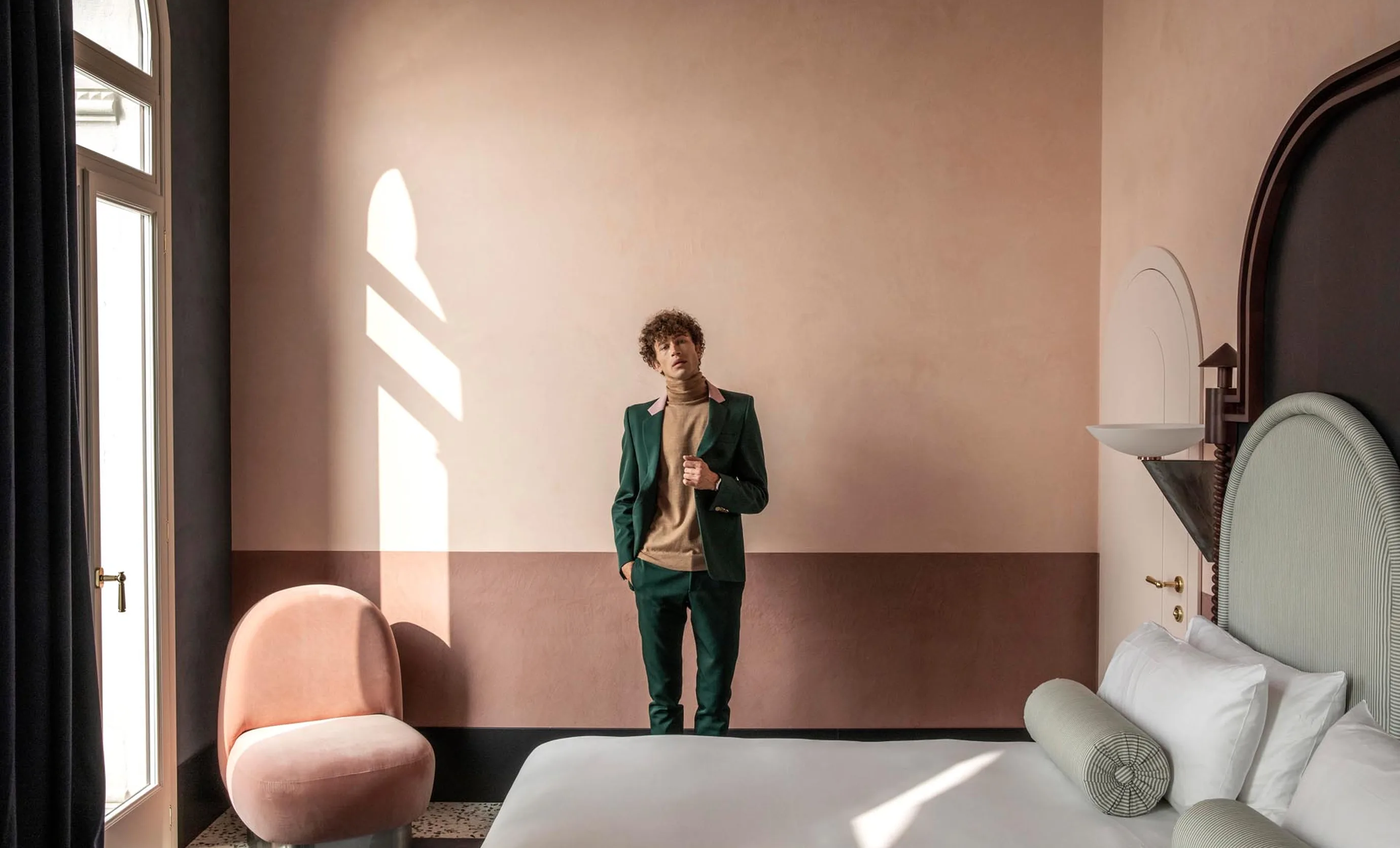 Man in green suit standing in a minimalist hotel room with a large bed and pink chair near window.