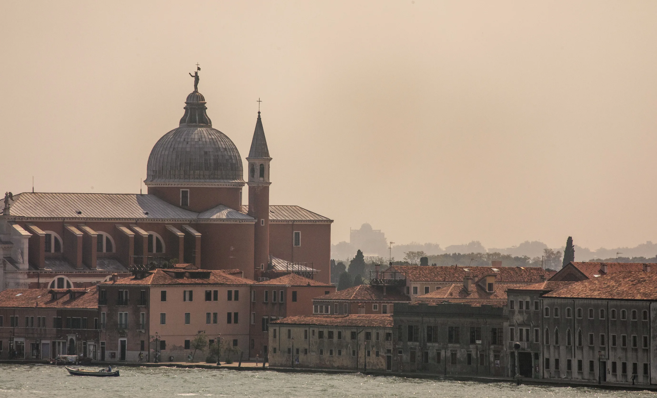 Historic buildings with red-tiled roofs along a waterfront under a hazy sky, featuring a large domed church and a smaller bell tower.