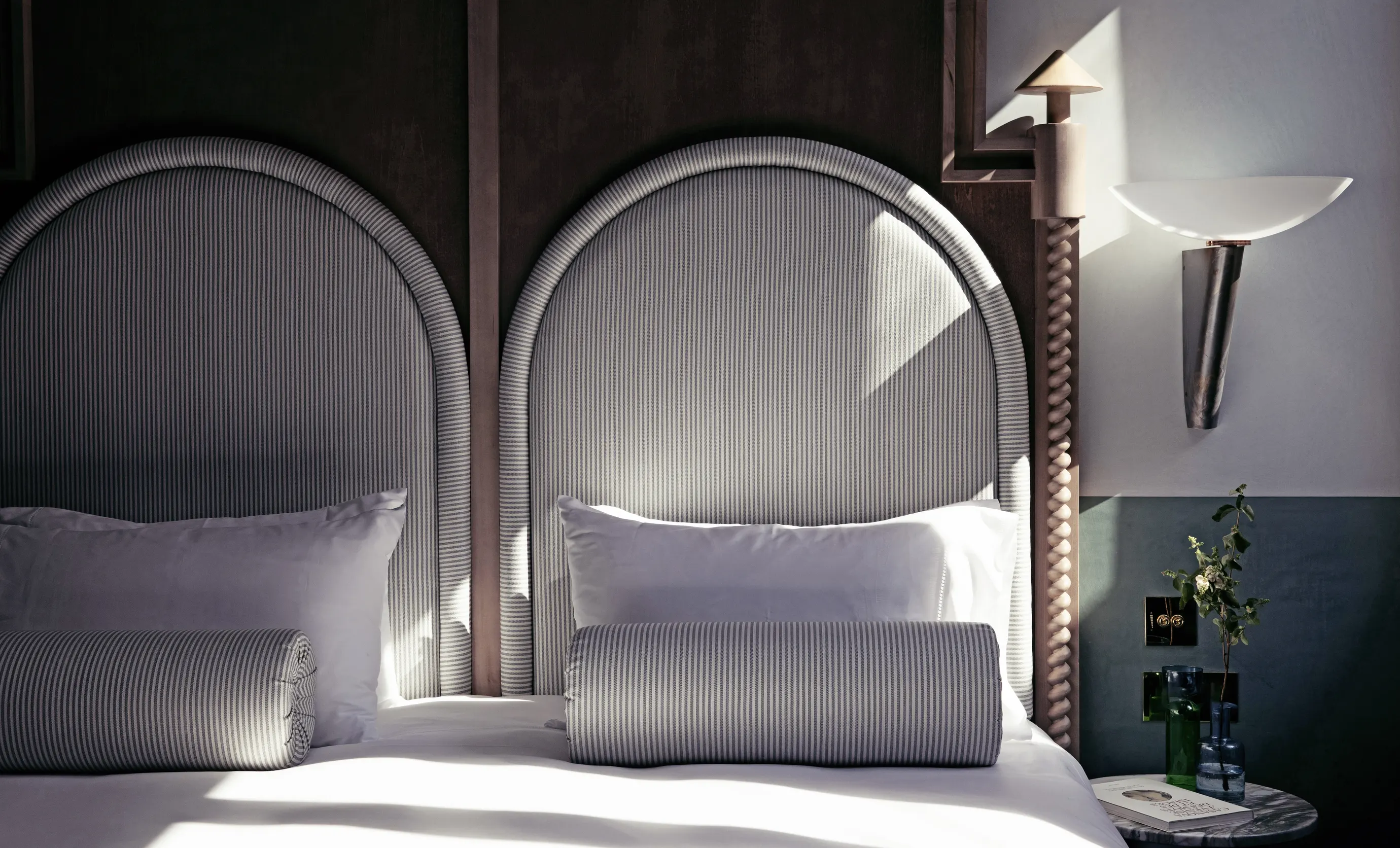 Hotel room bed with two rounded, vertically striped headboards, white pillows, striped cylindrical cushions, a wall sconce, and a small round side table with vases and a book.