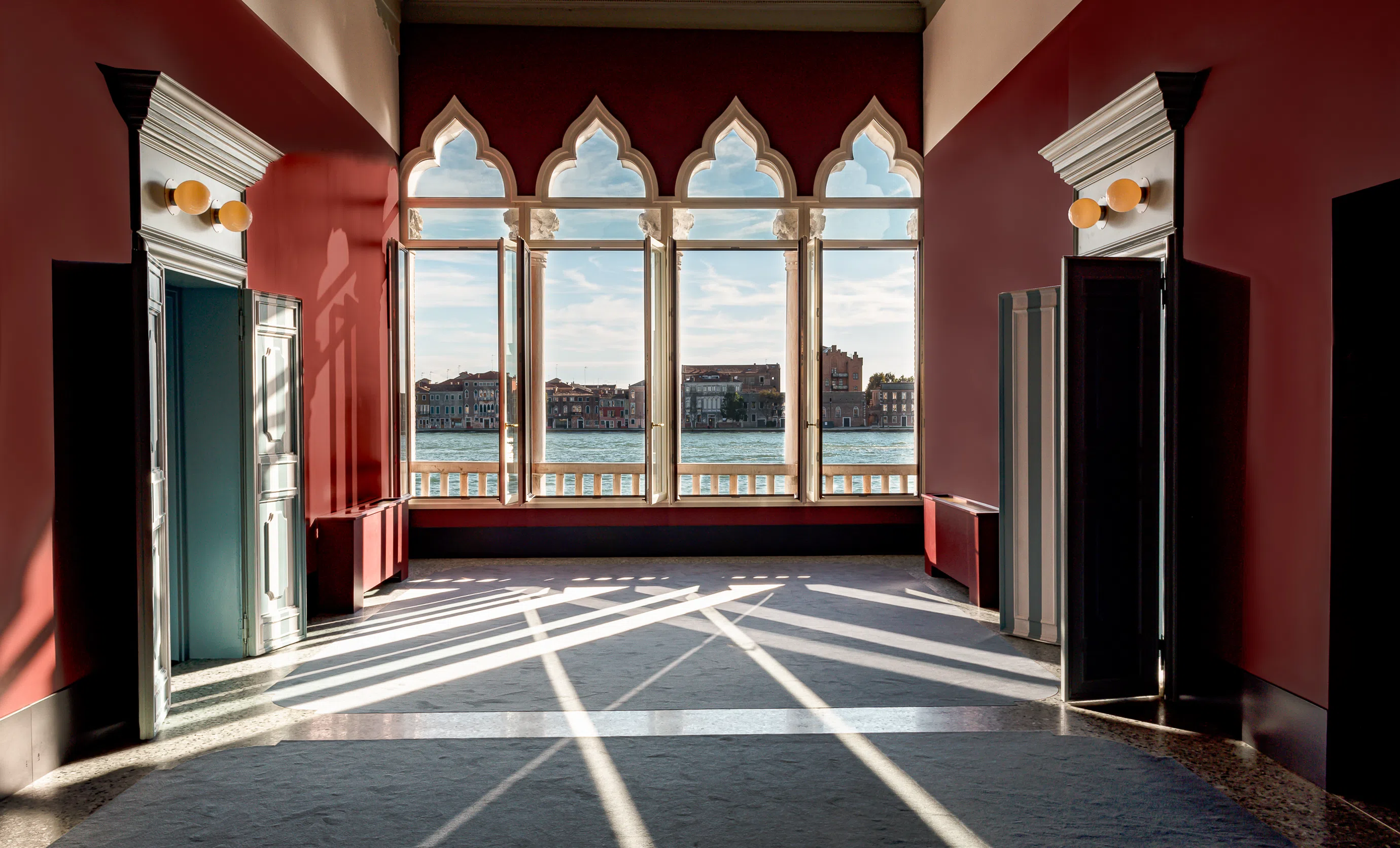 Room with red walls featuring Gothic-style windows overlooking water and distant buildings with sunlight casting shadows inside.