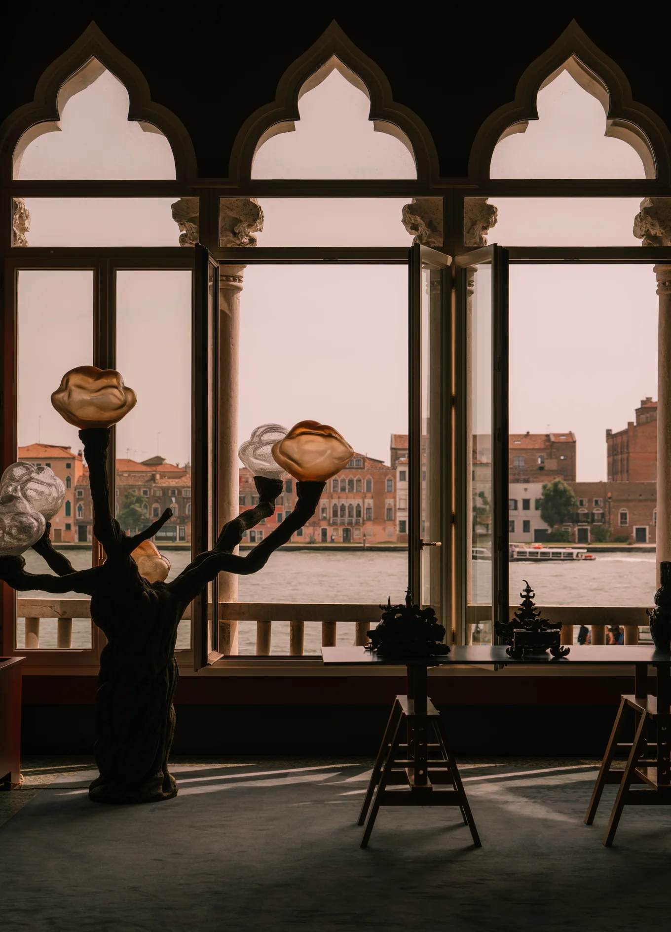 Interior view of a room with sculptural lamps shaped like flowers in front of large windows overlooking a canal and historic buildings outside.