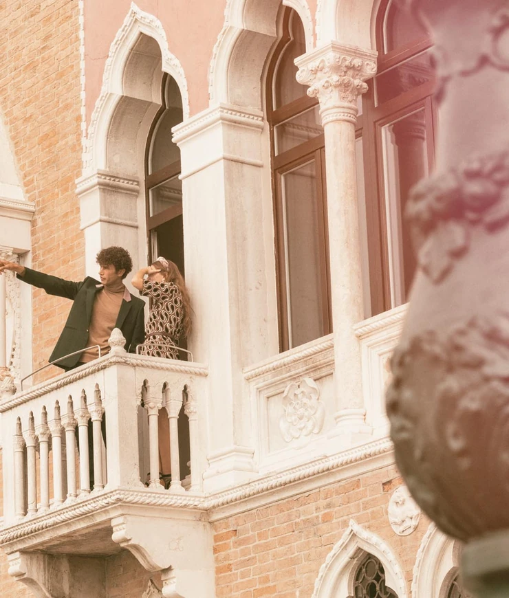 Man and woman standing on a balcony of a historic brick building with arched windows and ornate stone columns.
