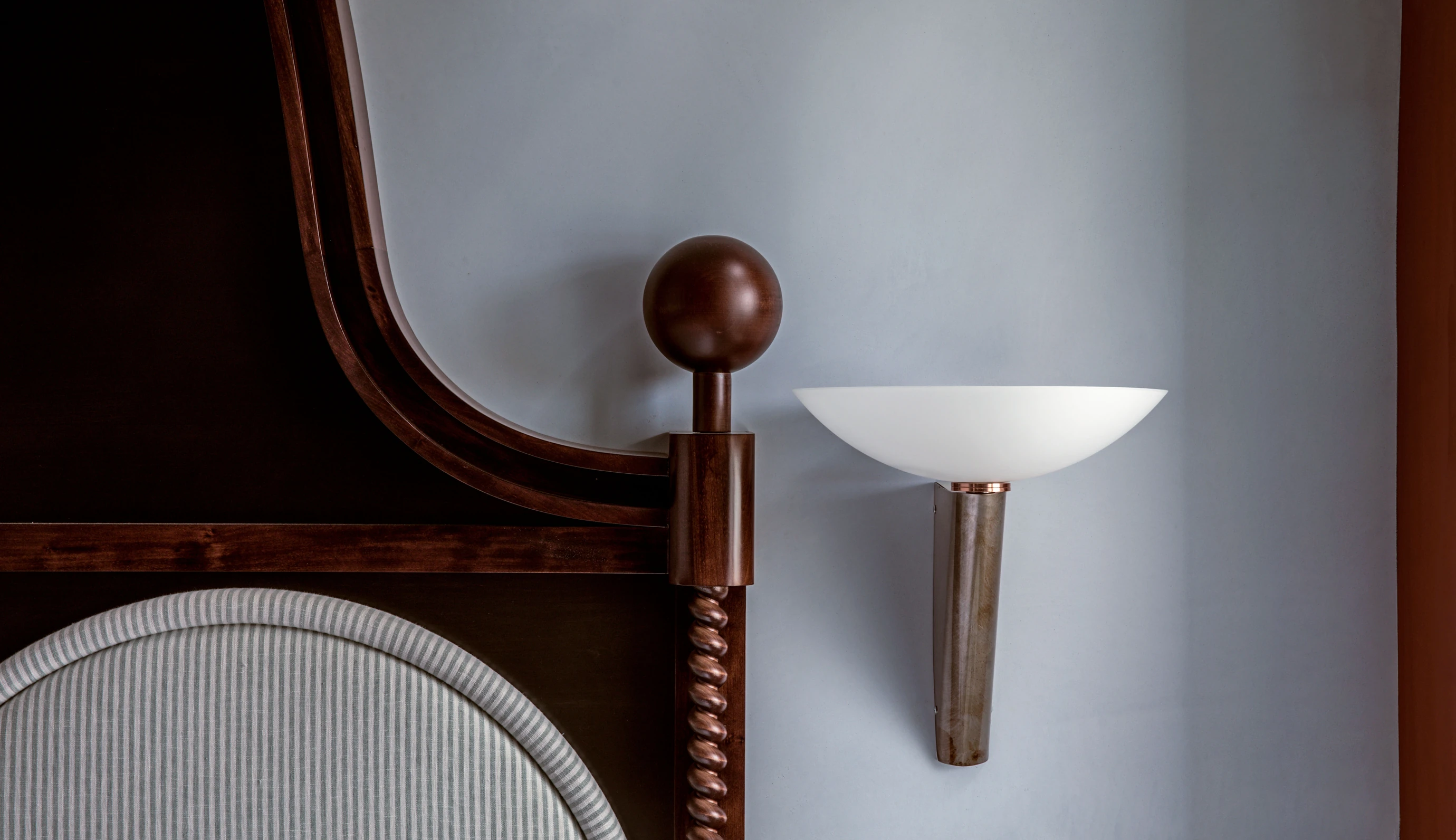 Close-up of an ornate wooden bedframe with a striped upholstered headboard next to a modern wall-mounted lamp with a white bowl-shaped shade.